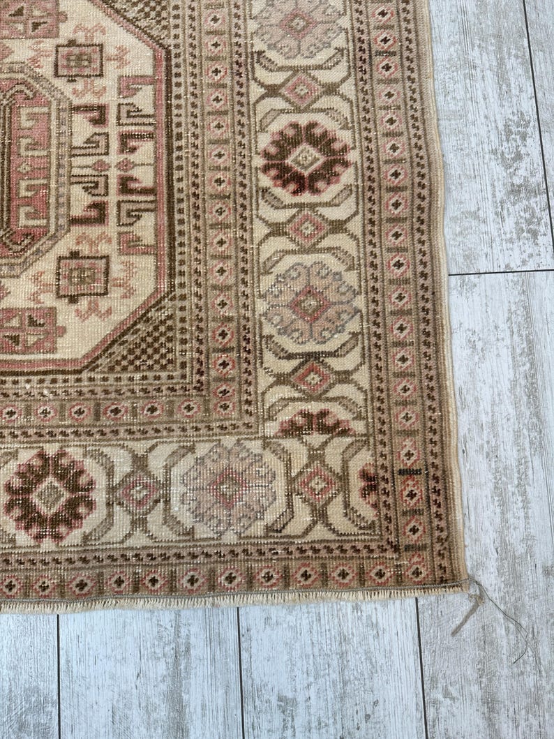 May include: Close-up of the bottom edge of a beige rug with a brown and pink geometric pattern. The rug has a fringe border.
