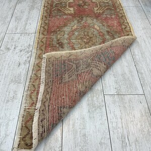 Puede incluir: Una peque&ntilde;a alfombra rectangular con un aspecto envejecido y vintage. La alfombra presenta un dise&ntilde;o central en tonos rojos, marrones y beige, con patrones geom&eacute;tricos. Los bordes est&aacute;n deshilachados y parte de la alfombra est&aacute; doblada, revelando la parte inferior.