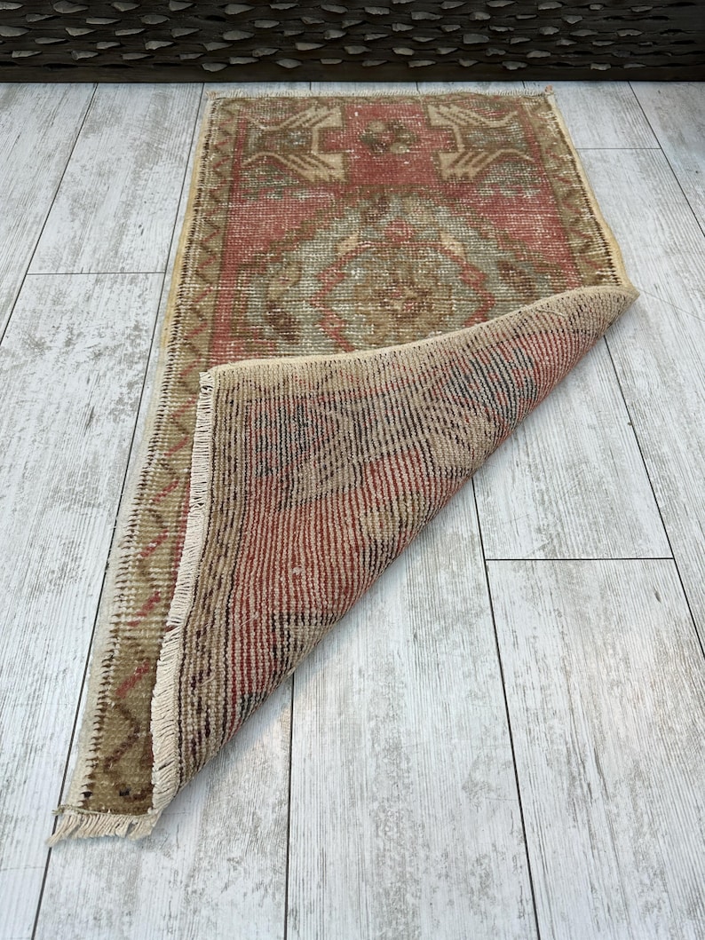 Puede incluir: Una peque&ntilde;a alfombra rectangular con un aspecto envejecido y vintage. La alfombra presenta un dise&ntilde;o central en tonos rojos, marrones y beige, con patrones geom&eacute;tricos. Los bordes est&aacute;n deshilachados y parte de la alfombra est&aacute; doblada, revelando la parte inferior.