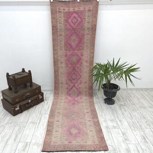 May include: A long, narrow runner rug featuring a faded beige and tan base with a repeating series of vibrant magenta diamond-shaped geometric patterns running down the center. The rug has a textured, woven appearance with a fringed edge at the top.