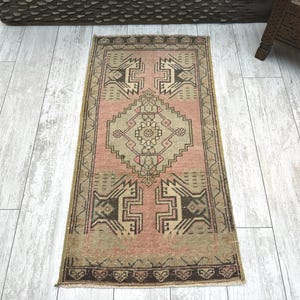 May include: A rectangular, vintage rug with a central geometric design in shades of pink, beige, and brown. The rug features a detailed border and intricate patterns, showcasing a traditional aesthetic. The rug is placed on a light-coloured wooden floor.