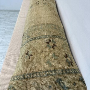 May include: A long, rectangular bolster cushion featuring a woven, textured surface in shades of beige, tan, and muted blue. The fabric displays a repeating geometric pattern with diamond shapes and small floral motifs across the entire length.