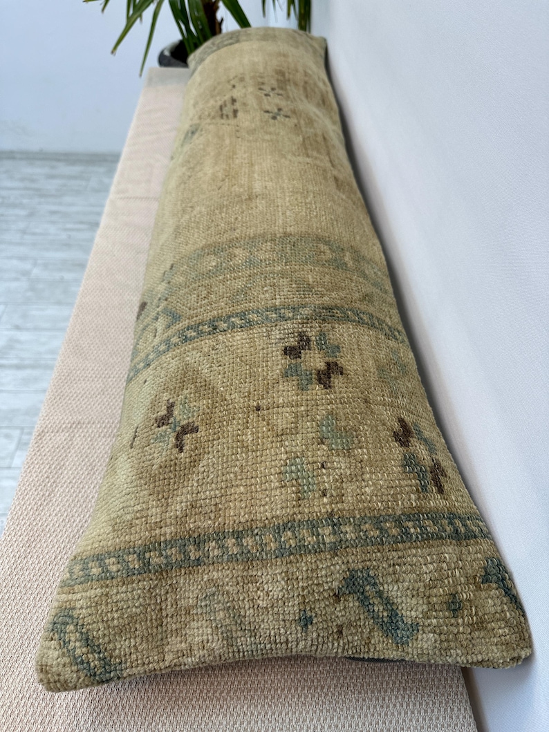 May include: A long, rectangular bolster cushion featuring a woven, textured surface in shades of beige, tan, and muted blue. The fabric displays a repeating geometric pattern with diamond shapes and small floral motifs across the entire length.