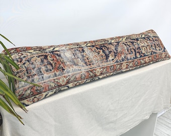 Puede incluir: Un cojín largo y rectangular con un diseño de alfombra vintage. El cojín presenta un patrón con tonos de azul marino, óxido y crema. El cojín está colocado sobre una superficie blanca.
