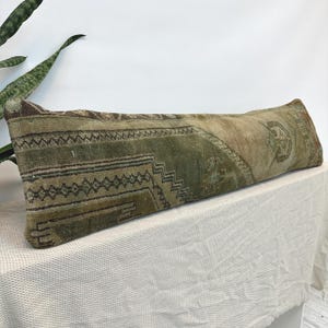 May include: A long, rectangular lumbar cushion featuring a faded, distressed rug-style pattern in shades of olive green, beige, and brown. The textile displays intricate geometric motifs and linear borders, showcasing a worn, textured appearance.