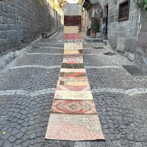 Puede incluir: Una alfombra larga y rectangular de patchwork compuesta por varias secciones estampadas en tonos rosa, beige y marrón. La alfombra se exhibe al aire libre en una calle adoquinada, mostrando su longitud y diseño.