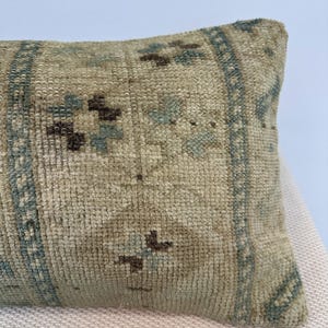 May include: A rectangular throw cushion featuring a textured, woven surface in a light beige tone. The fabric displays a repeating geometric pattern in muted shades of blue and dark brown, organised into vertical bands and central diamond motifs.