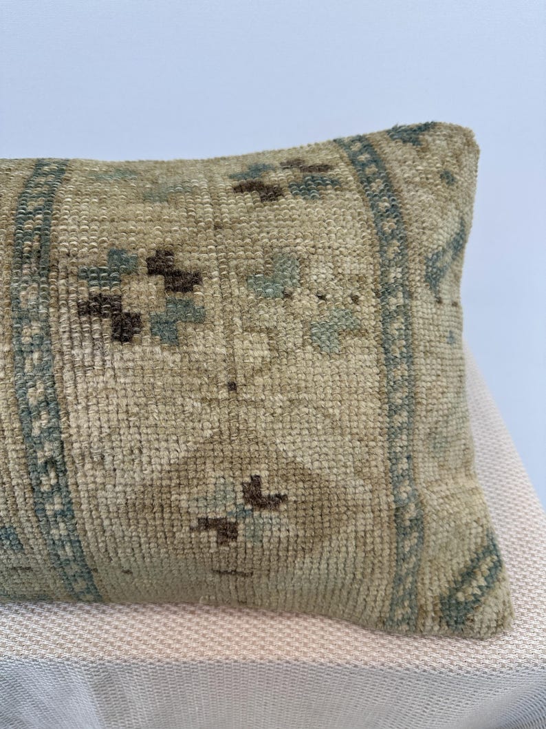 May include: A rectangular throw cushion featuring a textured, woven surface in a light beige tone. The fabric displays a repeating geometric pattern in muted shades of blue and dark brown, organised into vertical bands and central diamond motifs.