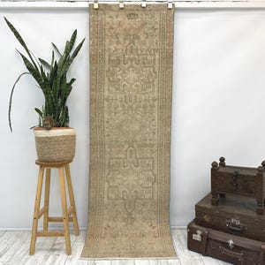 May include: A long, narrow runner rug featuring a muted beige and tan color palette with intricate geometric patterns and symmetrical motifs. The rug is displayed hanging vertically, showcasing a detailed woven texture and a fringed edge at the top.