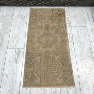 May include: A rectangular area rug featuring a muted tan and brown color palette with a central geometric medallion and symmetrical angular patterns throughout the woven surface, finished with short fringe along the top and bottom edges.