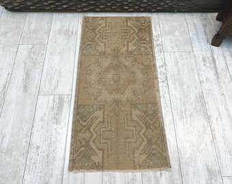 May include: A rectangular area rug featuring a muted tan and brown color palette with a central geometric medallion and symmetrical angular patterns throughout the woven surface, finished with short fringe along the top and bottom edges.