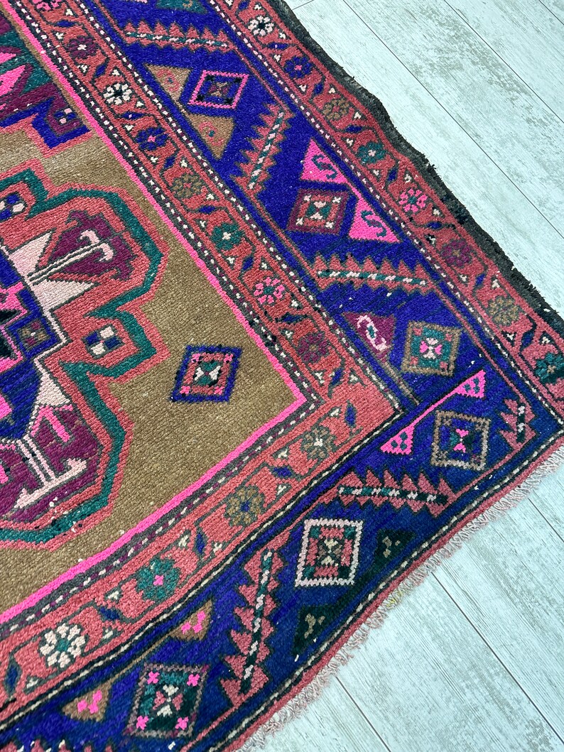 May include: A vintage rug with a brown background and a blue, pink, and green geometric pattern. The rug has a fringed edge.