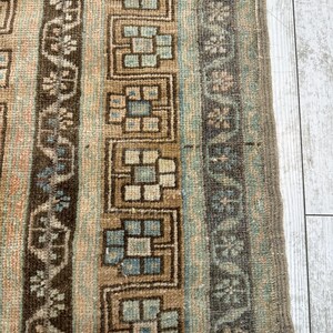May include: Close-up of a section of a vintage rug with a geometric pattern. The rug features vertical stripes in shades of brown, beige, and blue-green. The design includes floral and block-like motifs, with a fringed edge.