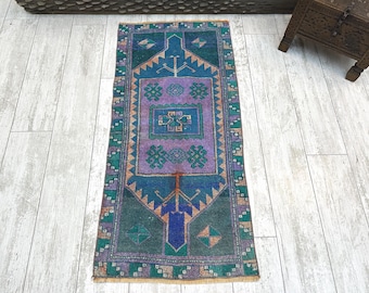 May include: A rectangular area rug featuring a central purple field with geometric motifs, surrounded by a dark blue and green border. The design includes intricate patterns in shades of teal, orange, and blue, with a fringed edge at both ends.