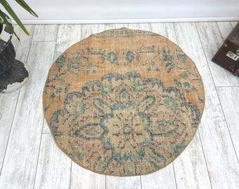 Puede incluir: Alfombra redonda de estilo vintage con un aspecto desgastado y descolorido. La alfombra presenta un diseño de medallón central en tonos turquesa y azul sobre un fondo cálido de color naranja melocotón. Los bordes de la alfombra están ligeramente desgastados, lo que aumenta su encanto antiguo.