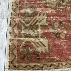 Puede incluir: Primer plano de una peque&ntilde;a alfombra rectangular con una est&eacute;tica vintage descolorida. La alfombra presenta un campo central de color rojo apagado con patrones geom&eacute;tricos beige y verde oliva. Los bordes est&aacute;n ribeteados con un borde beige y flecos.