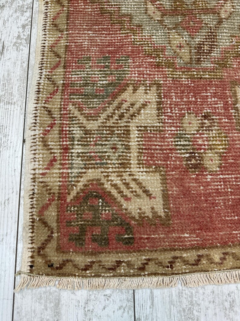 Puede incluir: Primer plano de una peque&ntilde;a alfombra rectangular con una est&eacute;tica vintage descolorida. La alfombra presenta un campo central de color rojo apagado con patrones geom&eacute;tricos beige y verde oliva. Los bordes est&aacute;n ribeteados con un borde beige y flecos.