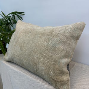May include: A rectangular throw cushion featuring a textured, woven surface in a neutral beige and tan colour palette. The fabric displays a subtle, weathered appearance with faint, faded patterns visible across the front of the cushion cover.
