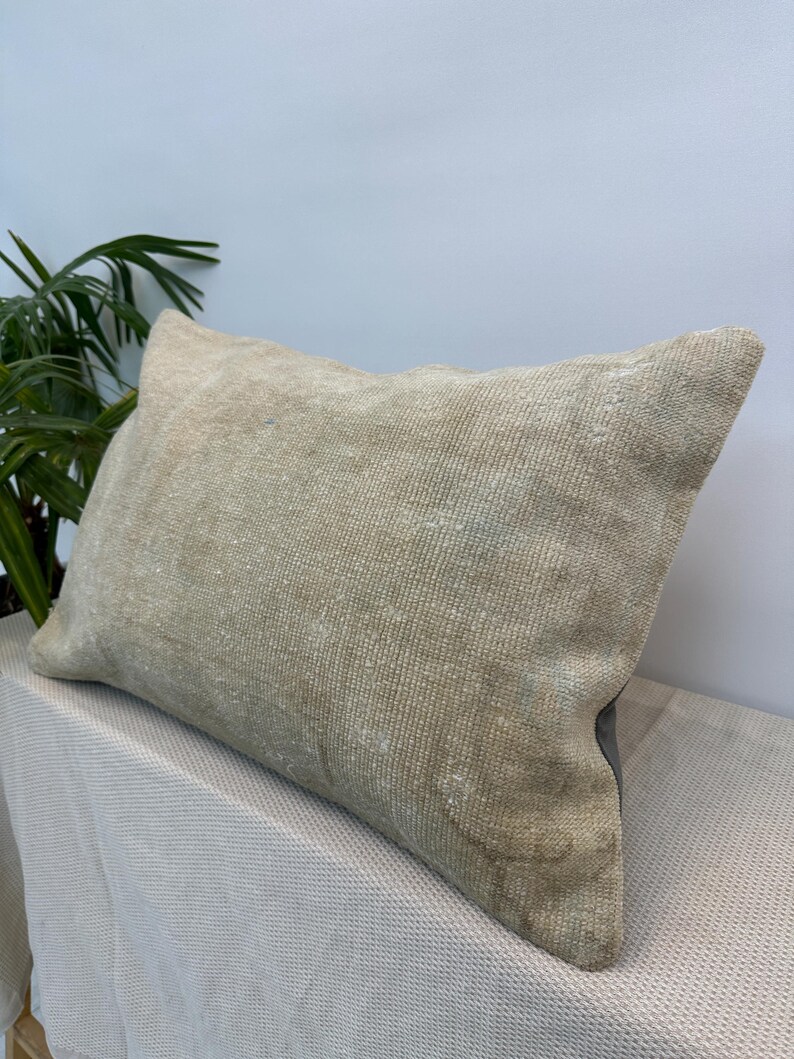 May include: A rectangular throw cushion featuring a textured, woven surface in a neutral beige and tan colour palette. The fabric displays a subtle, weathered appearance with faint, faded patterns visible across the front of the cushion cover.
