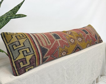 Puede incluir: Un cojín lumbar rectangular largo con un patrón textil tejido en tonos amarillo mostaza, rojo terracota, gris carbón y morado apagado. El diseño incluye bordes geométricos de grecas y motivos florales.