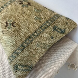 May include: A rectangular throw cushion featuring a woven texture in shades of beige, tan, and muted green. The surface displays a repeating geometric pattern with small dark brown and teal accents arranged in a structured, bordered design.
