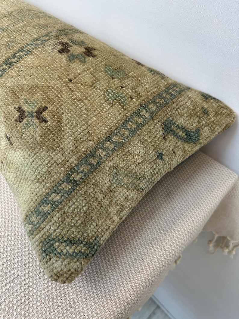 May include: A rectangular throw cushion featuring a woven texture in shades of beige, tan, and muted green. The surface displays a repeating geometric pattern with small dark brown and teal accents arranged in a structured, bordered design.