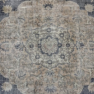 K&ouml;nnte beinhalten: Ein Vintage-Teppich mit einem verblassten, komplizierten Design in Blau- und Beiget&ouml;nen. Der Teppich hat ein zentrales Medaillon mit einem Blumenmuster und einen sich wiederholenden geometrischen Rand.