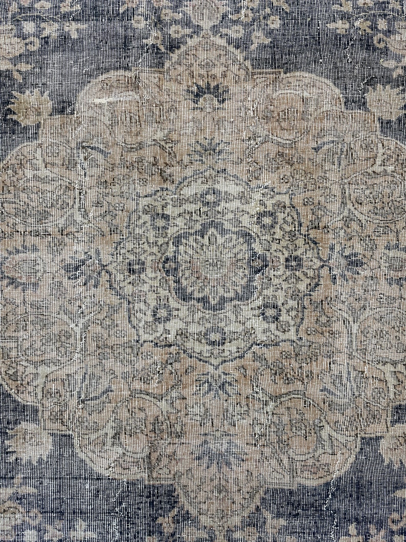 K&ouml;nnte beinhalten: Ein Vintage-Teppich mit einem verblassten, komplizierten Design in Blau- und Beiget&ouml;nen. Der Teppich hat ein zentrales Medaillon mit einem Blumenmuster und einen sich wiederholenden geometrischen Rand.