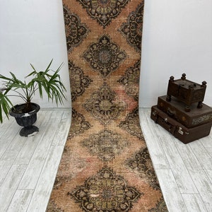 Puede incluir: Una alfombra de pasillo vintage con un fondo rosa descolorido y intrincados patrones florales negros y verdes. La alfombra tiene un aspecto desgastado y es perfecta para a&ntilde;adir un toque de estilo bohemio a cualquier habitaci&oacute;n.