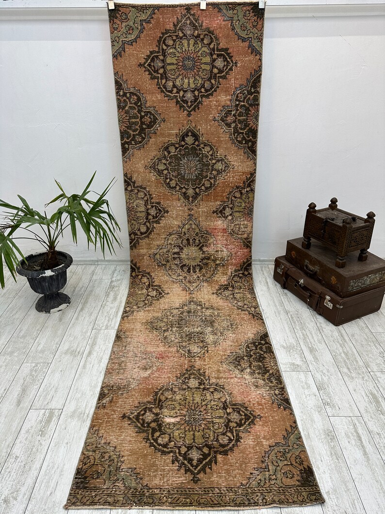 Puede incluir: Una alfombra de pasillo vintage con un fondo rosa descolorido y intrincados patrones florales negros y verdes. La alfombra tiene un aspecto desgastado y es perfecta para a&ntilde;adir un toque de estilo bohemio a cualquier habitaci&oacute;n.