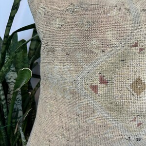 May include: A square throw pillow featuring a textured, woven surface in muted beige and cream tones. The fabric displays a faded geometric pattern with subtle accents of dusty rose and sage green, creating a soft, weathered appearance.