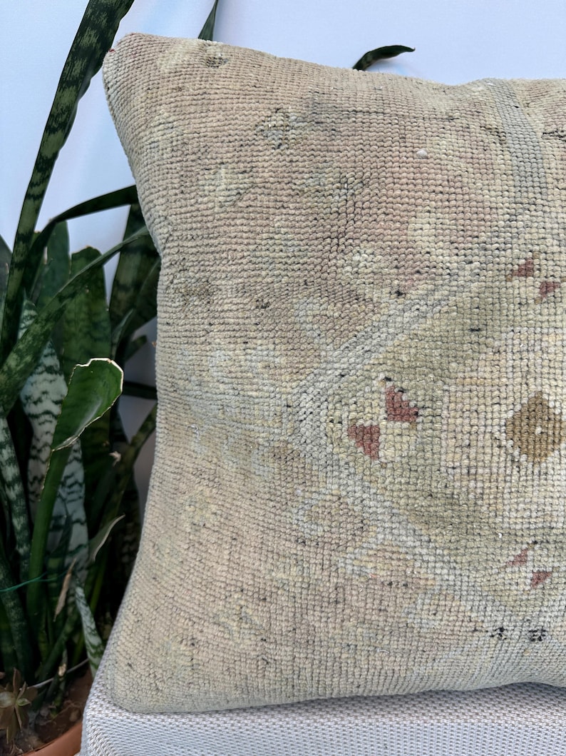 May include: A square throw pillow featuring a textured, woven surface in muted beige and cream tones. The fabric displays a faded geometric pattern with subtle accents of dusty rose and sage green, creating a soft, weathered appearance.