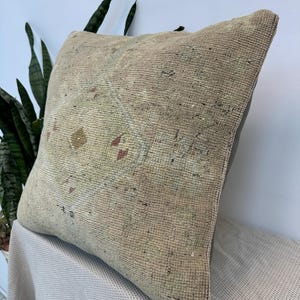 May include: A square throw pillow featuring a weathered, beige, and tan woven fabric with a faded, central geometric medallion pattern in muted tones of brown, yellow, and red. The pillow has a textured, coarse surface and a solid dark gray back.