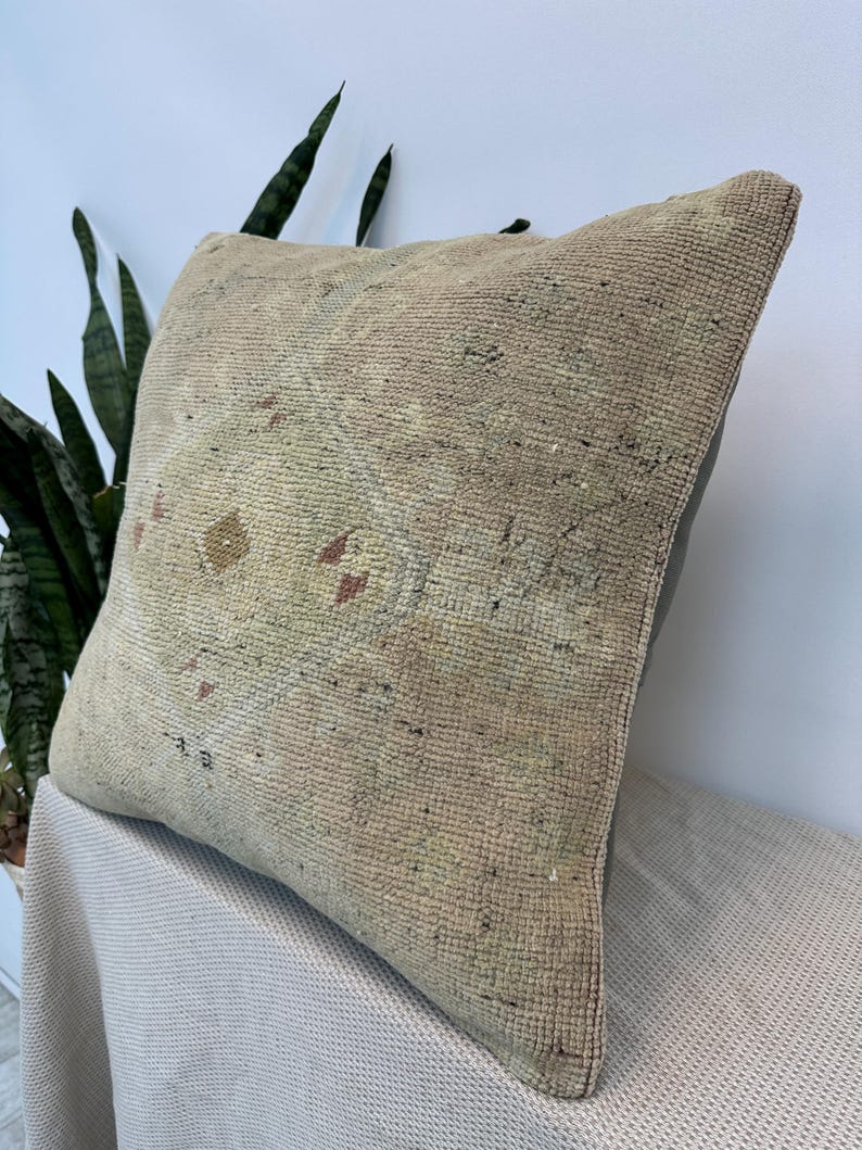 May include: A square throw pillow featuring a weathered, beige, and tan woven fabric with a faded, central geometric medallion pattern in muted tones of brown, yellow, and red. The pillow has a textured, coarse surface and a solid dark gray back.
