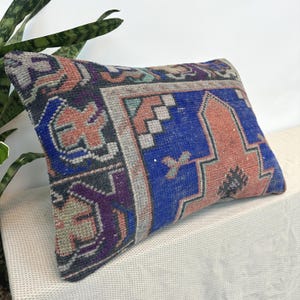 May include: A rectangular throw pillow featuring a woven textile cover with a geometric pattern in shades of deep blue, muted orange, purple, and grey. The design includes intricate borders and central motifs characteristic of a flat-woven rug.