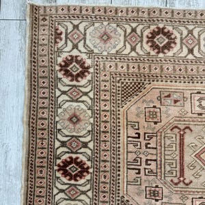 May include: A vintage rug with a cream background and intricate patterns in shades of brown, pink, and red. The rug features a central medallion with a geometric design and a border with repeating floral motifs.