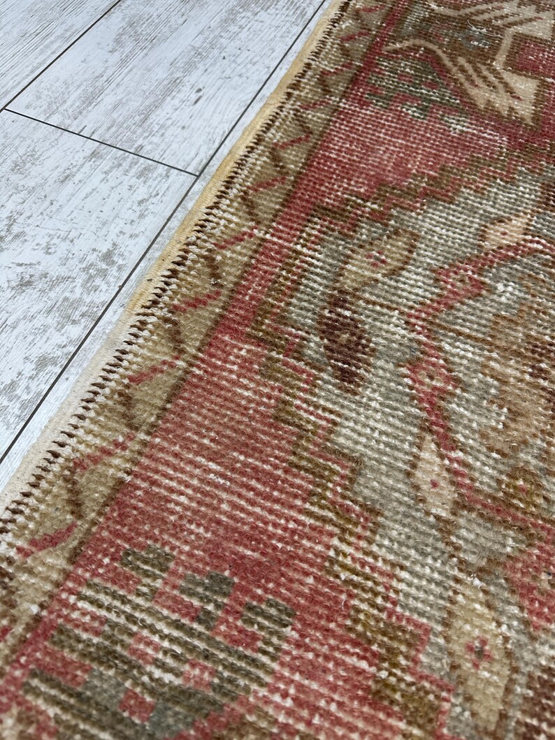 Puede incluir: Primer plano de una alfombra vintage con un aspecto desgastado y descolorido. La alfombra presenta una paleta de colores rojo, beige y marr&oacute;n, con intrincados patrones geom&eacute;tricos y una superficie texturizada. Los bordes de la alfombra est&aacute;n acabados con un borde cosido.