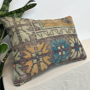 May include: A rectangular throw pillow featuring a woven textile pattern with a distressed appearance. The design includes geometric floral motifs in shades of mustard yellow, bright blue, dark brown, and muted sage green on a textured surface.