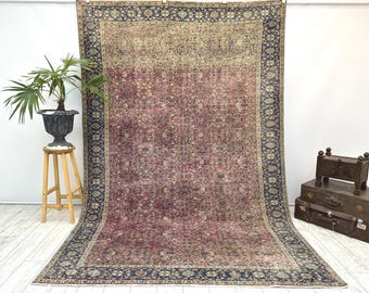 May include: A large, rectangular rug with a faded, vintage appearance. The rug features a central field of deep red with intricate floral patterns. A dark blue border with floral motifs frames the rug. The rug is displayed indoors.