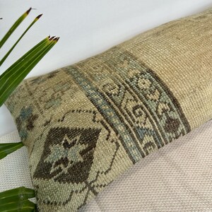 May include: A long rectangular lumbar cushion featuring a woven texture in shades of beige, tan, and light brown. The surface displays a central band with intricate geometric patterns in muted blue, dark brown, and cream, including diamond shapes.