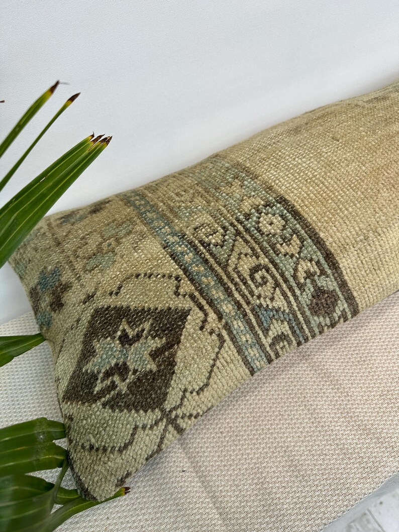 May include: A long rectangular lumbar cushion featuring a woven texture in shades of beige, tan, and light brown. The surface displays a central band with intricate geometric patterns in muted blue, dark brown, and cream, including diamond shapes.
