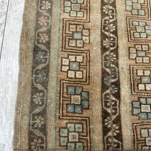 May include: Close-up of a section of a rug with a geometric pattern. The rug features vertical stripes in shades of brown, beige, and blue. The design includes repeating square and floral motifs, creating a textured appearance. The rug's edge is visible.