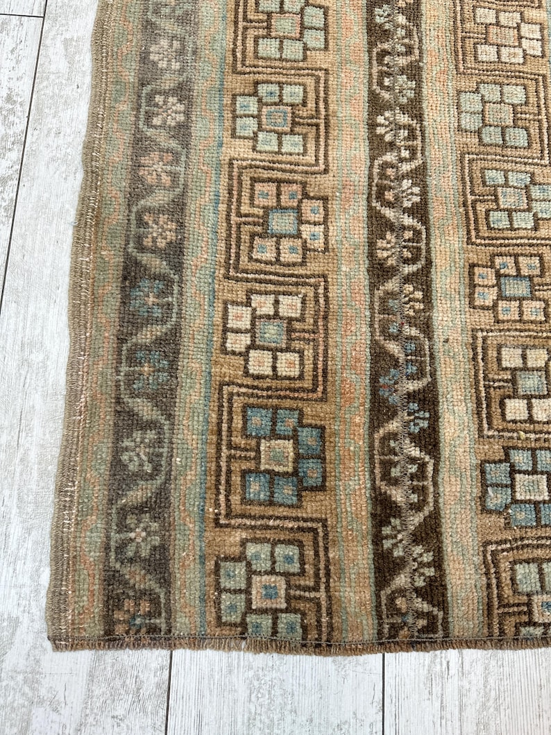 May include: Close-up of a section of a rug with a geometric pattern. The rug features vertical stripes in shades of brown, beige, and blue. The design includes repeating square and floral motifs, creating a textured appearance. The rug's edge is visible.