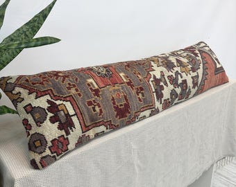 Puede incluir: Un cojín lumbar rectangular largo con una funda de tejido textil que presenta un complejo patrón geométrico en tonos terracota, rojo oscuro, gris carbón y crema. El diseño muestra intrincados motivos de inspiración tribal en la superficie.