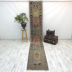 Puede incluir: Una alfombra de pasillo larga y estrecha con un patrón descolorido y multicolor en tonos rosa apagado, verde salvia, beige y negro carbón. El diseño incluye bordes geométricos intrincados y motivos florales centrales en una disposición vertical.