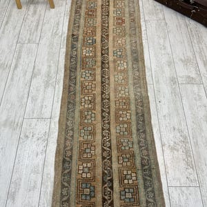 May include: A rectangular, vintage-style runner rug with a repeating geometric pattern in shades of brown, beige, and blue. The rug features vertical stripes with floral and square motifs. The rug is placed on a light-colored wooden floor.