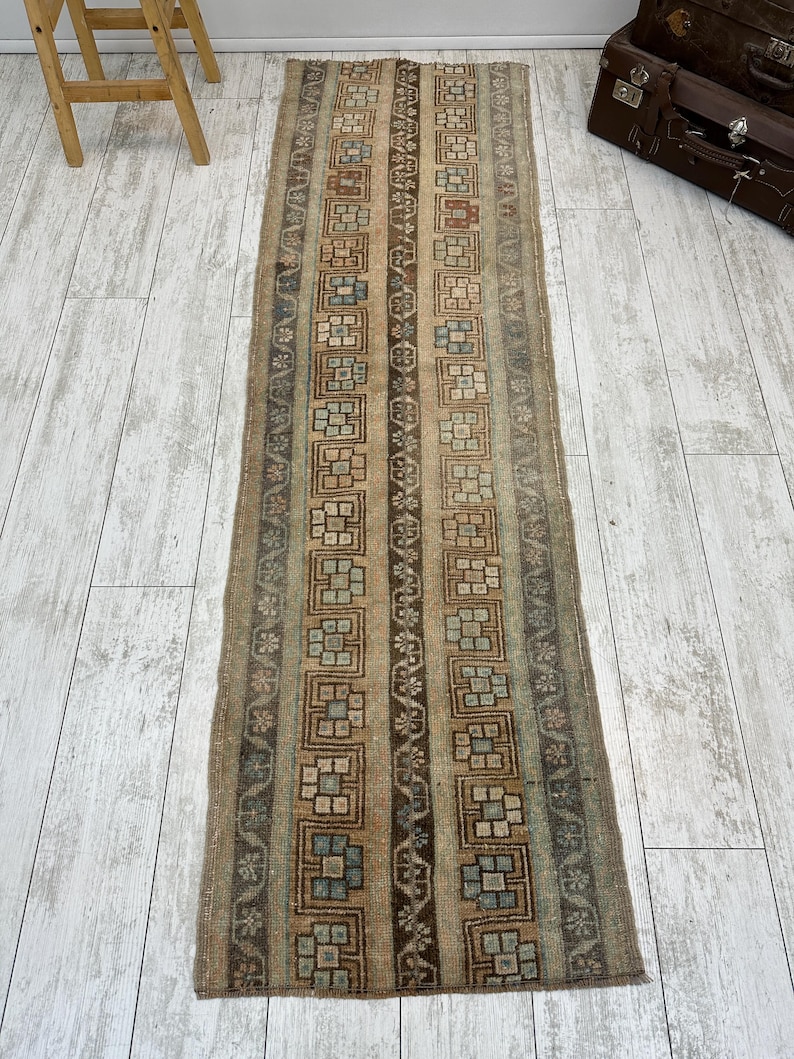 May include: A rectangular, vintage-style runner rug with a repeating geometric pattern in shades of brown, beige, and blue. The rug features vertical stripes with floral and square motifs. The rug is placed on a light-colored wooden floor.
