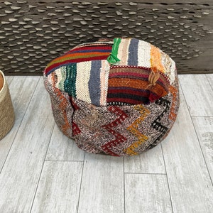 May include: A round, multi-colored, woven pouf with a patchwork design. The pouf is made of different colored fabrics, including red, orange, yellow, green, blue, and brown. The pouf is sitting on a light wood floor.