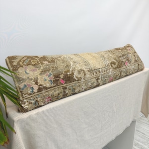 May include: A long, rectangular lumbar cushion featuring a faded, distressed pattern in shades of brown, beige, and cream. The textile displays intricate, worn-in floral and geometric motifs with small accents of muted pink and light blue throughout.