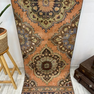 May include: A vintage Turkish runner rug with a faded peach background and intricate floral patterns in shades of green, brown, and blue. The rug has a fringed edge and a geometric border.
